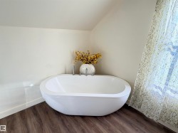 Freestanding oval soaking tub with deck-mounted fixtures, wood-finish flooring, and a large window featuring sheer patterned curtains - 