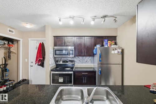 Kitchen featuring dark wood-finish cabinetry, stainless steel appliances, a patterned backsplash, and a black countertop with an integrated double basin sink - 424 1510 Watt Drive, Edmonton, AB 