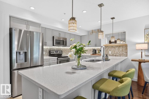 Kitchen featuring a spacious island with a light-toned countertop, stainless steel appliances, modern gray cabinetry, mosaic tile backsplash, and recessed lighting - 207 5025 Edgemont Boulevard, Edmonton, AB 