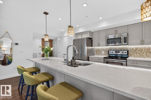 Spacious kitchen featuring a large island with a composite countertop, stainless steel appliances, light gray cabinetry, under-cabinet lighting, and a mosaic tile backsplash - 207 5025 Edgemont Boulevard, Edmonton, AB 