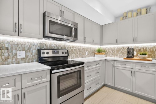 Contemporary kitchen featuring light gray cabinetry with chrome hardware, a patterned tile backsplash, under-cabinet lighting, stainless steel appliances, and light-toned countertops - 207 5025 Edgemont Boulevard, Edmonton, AB 