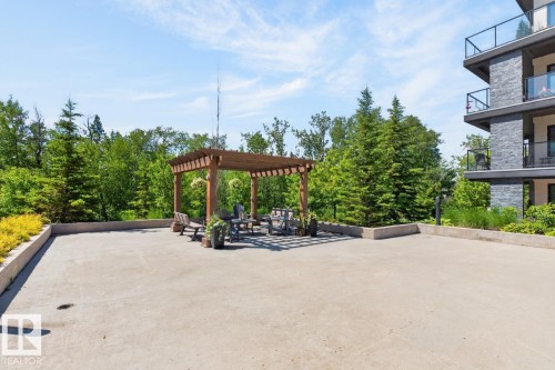 Expansive outdoor patio featuring a wooden pergola, concrete surfacing, raised planters with mature greenery, and a building exterior with stacked stone accents and glass-paneled balconies - 207 5025 Edgemont Boulevard, Edmonton, AB 
