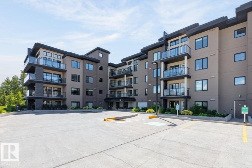 Contemporary condominium building featuring a multi-tone facade, ample parking, multiple private balconies with glass railings, modern exterior lighting, and landscaped grounds - 207 5025 Edgemont Boulevard, Edmonton, AB 