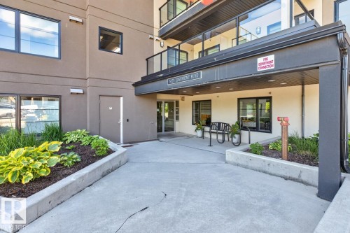 Building entry featuring covered portico, concrete landscaping beds, and established plantings - 207 5025 Edgemont Boulevard, Edmonton, AB 