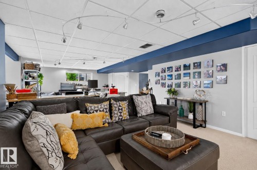 Spacious lower-level living area featuring a drop ceiling with track lighting, contrasting blue ceiling beams, light-toned carpeting, and a visible window for natural light - 163 Galland Crescent, Edmonton, AB 