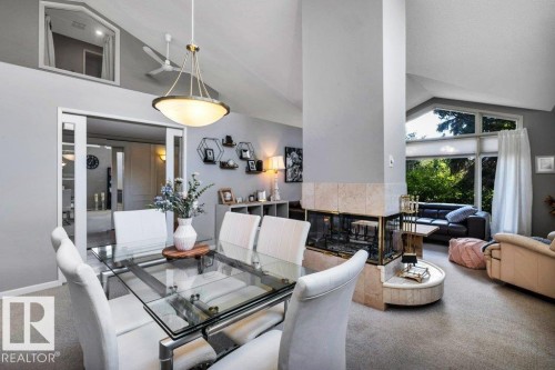 Vaulted ceiling dining area featuring a glass-top table, a central brass chandelier, a three-sided fireplace with a stone surround, an integrated stone hearth, and expansive windows - 11742 29 Avenue, Edmonton, AB 