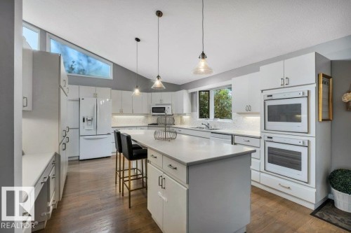 Modern kitchen featuring a central island with countertop seating, white cabinetry, integrated double ovens, wood-finish flooring, and a unique high-angle window - 11742 29 Avenue, Edmonton, AB 