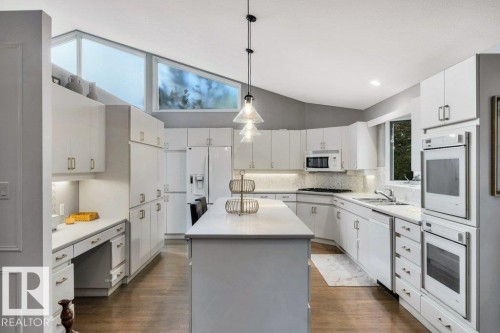 Kitchen featuring white cabinetry, a central island with pendant lighting, integrated built-in ovens, a desk area, and wood-finish flooring - 11742 29 Avenue, Edmonton, AB 