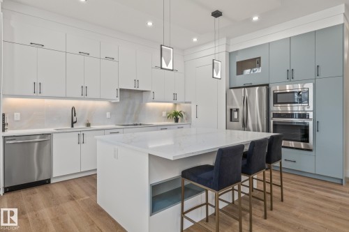 Gourmet kitchen featuring a large central island with seating, stainless steel appliances, white flat-panel cabinetry, a light-toned backsplash, and wood-finish flooring - 132 Edgewater Circle, Leduc, AB 