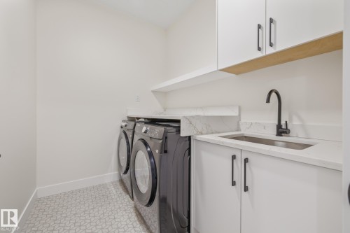 Dedicated laundry space featuring patterned floor tile, stone countertops, a stainless steel sink with a matte black faucet, and white cabinetry with dark hardware - 132 Edgewater Circle, Leduc, AB 