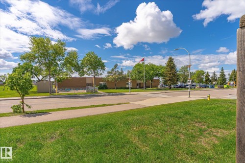 Grassy expanse with paved sidewalk and street, featuring mature trees and a distant building with a Canadian flag - 186 Cornell Crest, Edmonton, AB 
