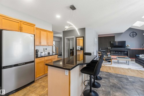 Open-concept kitchen and living area featuring a kitchen island with seating, stainless steel refrigerator, light wood cabinetry, black countertops, and a skylight in the living area - 3061 Spence Wynd, Edmonton, AB 