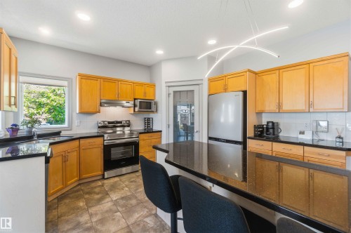 Contemporary kitchen featuring wood cabinetry, stainless steel appliances, dark countertops, a breakfast bar, and tile flooring - 3061 Spence Wynd, Edmonton, AB 
