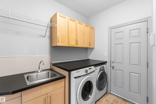 Dedicated laundry area featuring a stainless steel sink, black countertops, wood-finish cabinetry, and overhead wire shelving - 3061 Spence Wynd, Edmonton, AB 