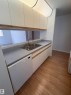 Kitchen area featuring a double basin stainless steel sink, a white built-in dishwasher, light wood-finish flooring, white cabinetry with wood trim, and a pass-through opening - 213 17467 98A Avenue, Edmonton, AB 