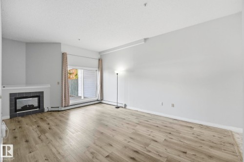 Spacious living area featuring wood-finish flooring, a corner fireplace with a dark tile surround, and direct access to an outdoor space through sliding glass doors - 105 11446 40 Avenue Nw, Edmonton, AB 