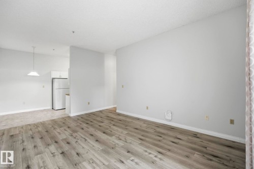 Spacious interior featuring wood-finish flooring, light-colored walls, and a recessed kitchen area with a white refrigerator and upper cabinetry - 105 11446 40 Avenue Nw, Edmonton, AB 