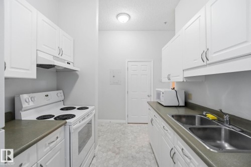 Galley kitchen featuring white cabinetry with silver-tone hardware, dark countertops, hexagonal floor tiling, a double basin stainless steel sink, and a white range with overhead ventilation - 105 11446 40 Avenue Nw, Edmonton, AB 