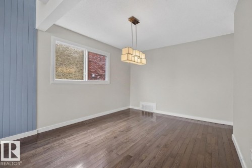 Spacious interior room featuring wood-finish flooring, a modern geometric chandelier, a window with white trim, and a prominent blue vertical plank accent wall - 2712 104 Street, Edmonton, AB 