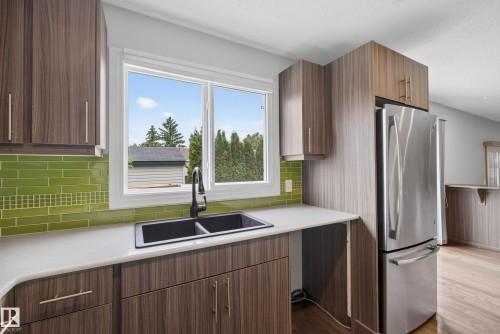 Kitchen featuring wood-grain cabinetry, a double-basin sink with matte black faucet, light-toned countertops, green subway tile backsplash, and stainless steel refrigerator - 2712 104 Street, Edmonton, AB 