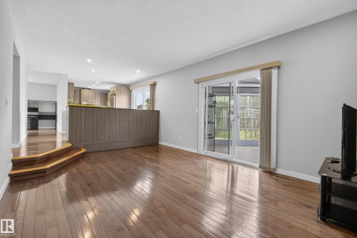 Open-concept living area featuring rich wood-finish flooring, a contemporary wood-paneled breakfast bar, a modern kitchen with stainless steel appliances, and sliding glass doors leading to an enclosed patio - 2712 104 Street, Edmonton, AB 