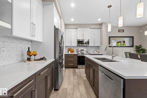 Contemporary kitchen featuring white shaker upper cabinetry and dark wood-finish lower cabinetry, a spacious island with a built-in stainless steel double sink and integrated dishwasher, stainless steel appliances, and wood-finish flooring - 2146 Koshal Way, Edmonton, AB 