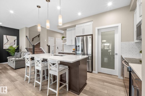 Open-concept kitchen featuring a large island with seating, stainless steel appliances, white shaker cabinetry, and a geometric tile backsplash - 2146 Koshal Way, Edmonton, AB 