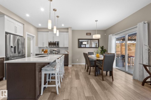 Open-concept kitchen and dining area featuring light wood-finish flooring, a kitchen island with seating, stainless steel appliances, white upper cabinetry, and a sliding glass door - 2146 Koshal Way, Edmonton, AB 