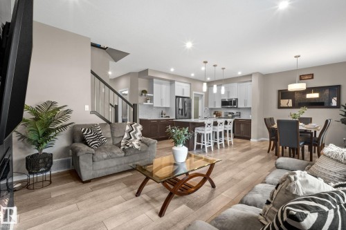 Open-concept living space featuring wood-finish flooring, a contemporary kitchen with two-tone cabinetry, stainless steel appliances, a kitchen island with seating, and a dining area with a modern chandelier - 2146 Koshal Way, Edmonton, AB 