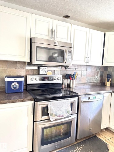 Kitchen featuring white cabinetry with silver-tone hardware, stainless steel appliances including an oven with a double door, a built-in microwave, and a dishwasher, complemented by a tiled backsplash and dark countertops - 45 Lambert Crescent, St. Albert, AB 