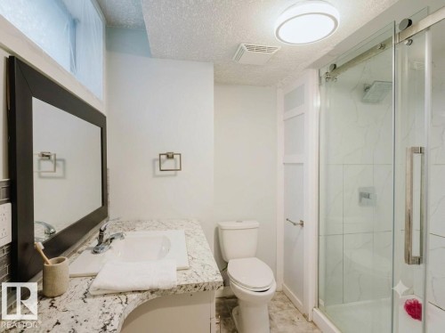 Bathroom featuring a granite countertop vanity with an undermount sink, a framed mirror, a toilet, and a glass-enclosed shower with tile surround and rain showerhead - 7515 130 Avenue, Edmonton, AB 