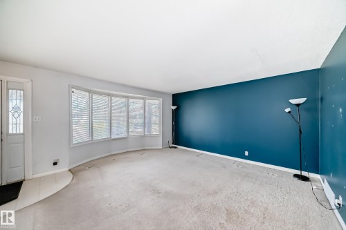 Spacious living area featuring a large bay window with horizontal blinds, a white entry door with decorative glass, and a distinct accent wall - 7317 83 Avenue, Edmonton, AB 