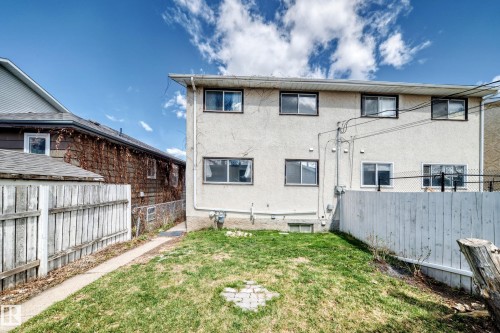 Rear exterior of the property featuring a two-story stucco facade, multiple windows with dark trim, white wood fencing, a partial chain-link fence, and a grassy yard space - 7317 83 Avenue, Edmonton, AB 