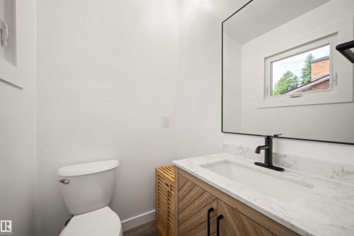 Bathroom featuring a single vanity with a white countertop and integrated sink, a wood-finish cabinet with black hardware, a rectangular mirror, a white toilet, and a white-framed window - 28 Goodridge Drive, St. Albert, AB 