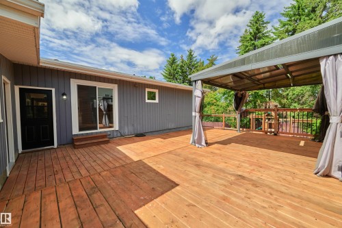 Expansive wood-finish deck featuring a covered pergola with privacy drapes, a sliding glass door entry, a separate solid black entry door, and a wooden railing - 28 Goodridge Drive, St. Albert, AB 