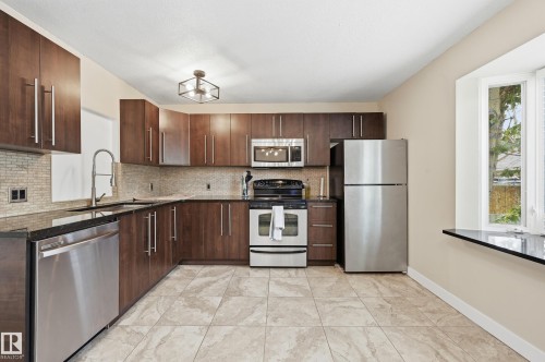 Kitchen featuring dark wood-finish cabinetry with brushed nickel hardware, stainless steel appliances, a mosaic tile backsplash, and dark countertops - 12326 92 Street, Edmonton, AB - Indoor Photo Showing Kitchen