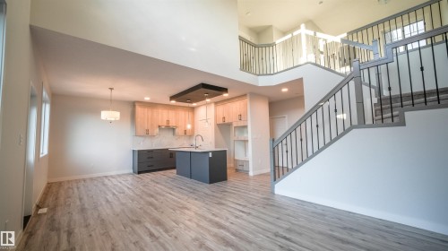 Open-concept living space featuring wood-finish flooring, a contemporary kitchen with a central island and recessed lighting, and a grand staircase with wrought iron balusters leading to a second-story landing - 44 Dalquist Bay, Leduc, AB 