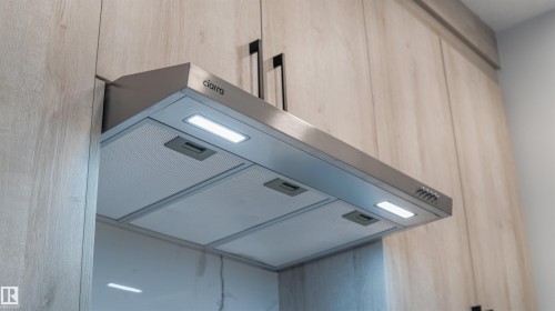 Stainless steel range hood with integrated LED lighting and button controls, framed by wood-finish cabinetry featuring matte black hardware - 44 Dalquist Bay, Leduc, AB 