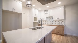 Kitchen featuring a large central island with an undermount double sink, white quartz countertops, sleek white upper cabinetry, natural wood-finish lower cabinetry, and a patterned backsplash - 