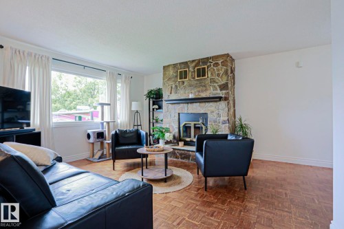 Living space featuring a stone-clad fireplace with a dark mantel, wood-finish parquet flooring, and a large window with white curtains - 2007 67 Street, Edmonton, AB 
