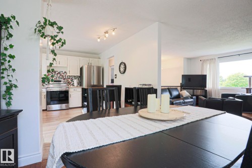 Open-concept living space featuring a black dining table, light wood-finish flooring, white kitchen cabinetry, stainless steel appliances, and a large window - 2007 67 Street, Edmonton, AB 