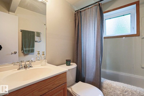 Bathroom featuring a wood-finish vanity with an integrated sink and chrome faucet, a white toilet, and a bathtub with a frosted window - 2007 67 Street, Edmonton, AB 