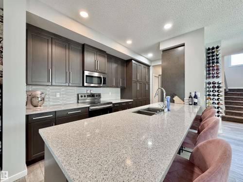 Kitchen featuring an extended island with a built-in sink, dark wood-finish cabinetry, stainless steel appliances, a textured backsplash, and recessed lighting - 302 Balsam Link, Leduc, AB 