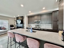 Kitchen featuring a large island with an integrated sink, dark wood-finish cabinetry, stainless steel appliances, a textured tile backsplash, and recessed lighting - 