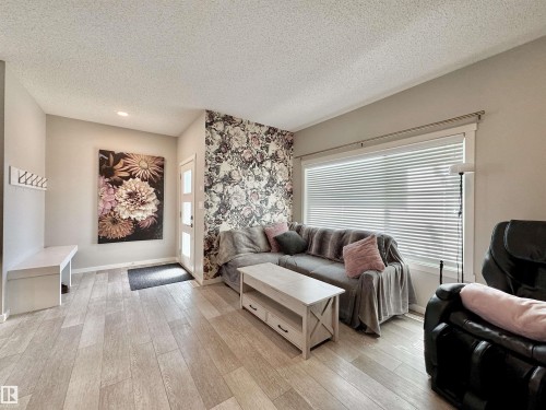 Spacious living area featuring light wood-finish flooring, a contemporary floral accent wall, and recessed ceiling lighting - 302 Balsam Link, Leduc, AB 