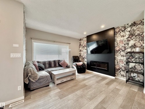 Living area featuring wood-finish flooring, recessed lighting, and a prominent feature wall with floral wallpaper and a dark-paneled fireplace surround - 302 Balsam Link, Leduc, AB 