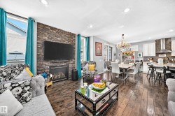 Open-concept living area featuring rich wood-finish flooring, a stone-accented fireplace wall, recessed lighting, a modern chandelier, and a kitchen with white cabinetry and a stainless steel range hood - 