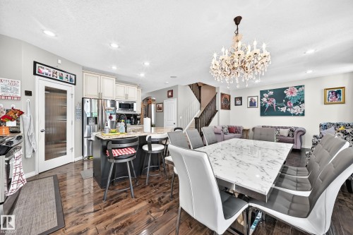 Open-concept living space featuring wood-finish flooring, a kitchen island with seating, stainless steel appliances, a crystal chandelier, and a staircase with dark wood railings - 12410 171A Avenue, Edmonton, AB 