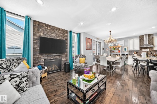 Open-concept living area featuring rich wood-finish flooring, a stone-accented fireplace wall, recessed lighting, a modern chandelier, and a kitchen with white cabinetry and a stainless steel range hood - 12410 171A Avenue, Edmonton, AB 