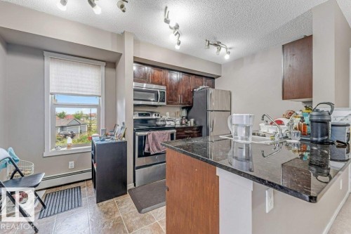 Kitchen featuring wood cabinetry, stainless steel appliances, a black stone countertop island, and tile flooring - 225 111 Watt Common, Edmonton, AB 